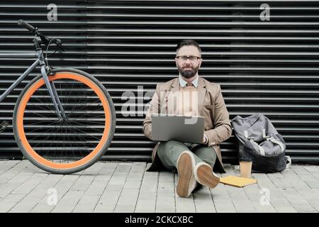 Portrait of mature bearded businessman sitting on the street and looking at camera while working on laptop Stock Photo