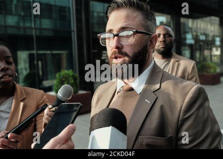 Mature bearded man in eyeglasses standing on the street among journalist and giving an interview Stock Photo