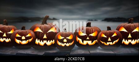 Seven spooky halloween pumpkins, Jack O Lantern, with an evil face and eyes on a wooden bench, table with a misty gray misty coastal night background. Stock Photo