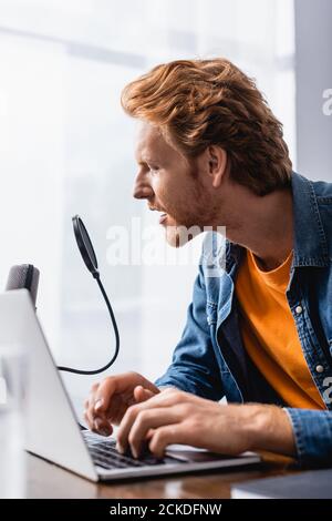 focused, redhead broadcaster speaking in microphone while using laptop Stock Photo