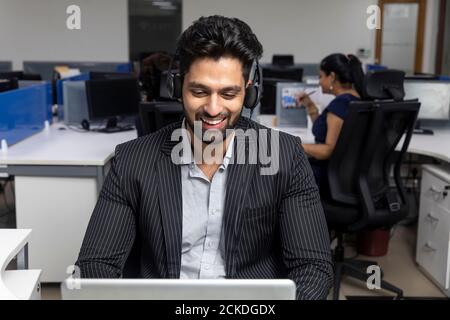 Portrait of young Indian businessman wearing headphones, sitting at his workstation against office background, corporate environment, call centre. Stock Photo