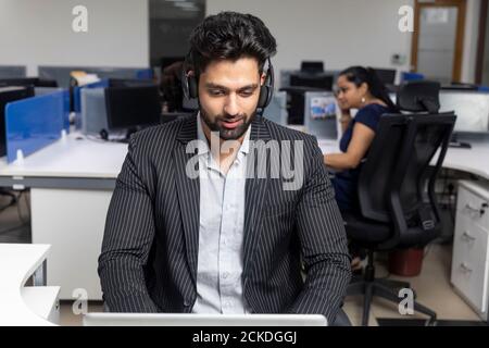 Portrait of young Indian businessman wearing headphones, sitting at his workstation against office background, corporate environment, call centre. Stock Photo