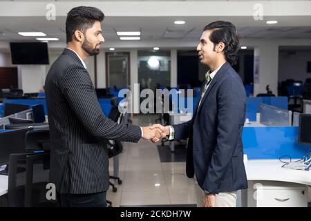 Portrait of two young Indian businessmen shaking hands, reaching agreement with client, closing a deal, corporate environment, office background. Stock Photo