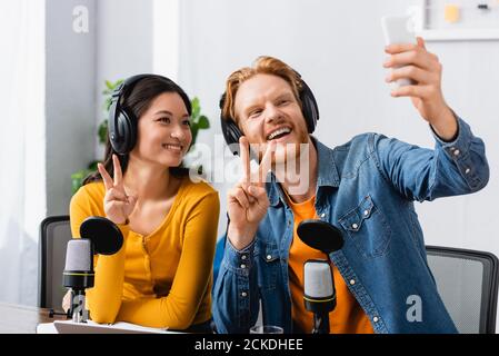excited, redhead announcer taking selfie on mobile phone while showing victory signs together with asian colleague Stock Photo