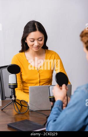 selective focus of young asian radio host using laptop during interview with man in studio Stock Photo