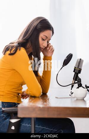 upset asian radio host touching face while sitting near microphone and cup of coffee with bowed head Stock Photo