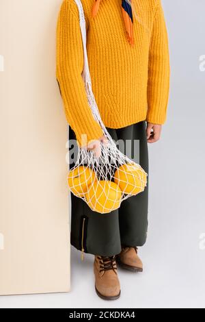 cropped view of fashionable girl in autumn outfit posing with grapefruits in string bag on beige and white background Stock Photo