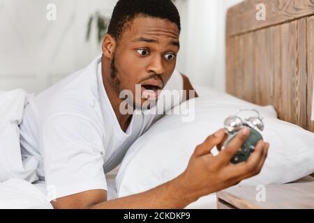 Overslept African Man Holding Alarm-Clock Waking Up In Bed Indoor Stock Photo