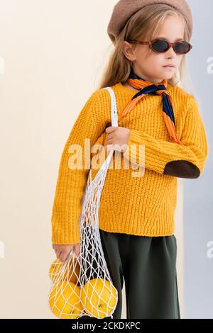 fashionable blonde girl in autumn outfit and sunglasses posing with grapefruits in string bag on beige and white background Stock Photo