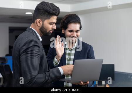 Portrait of two young handsome Indian businessmen waving hand at laptop, video conference call with client, discussing project, corporate environment. Stock Photo