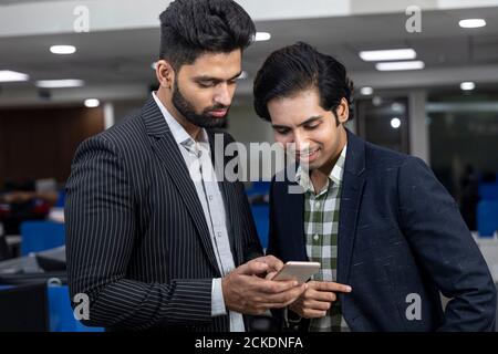 Portrait of two young handsome Indian businessmen looking into phone, discussing project, corporate environment. Stock Photo