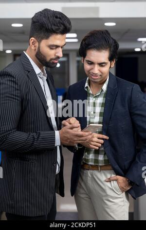 Portrait of two young handsome Indian businessmen looking into phone, discussing project, corporate environment. Stock Photo