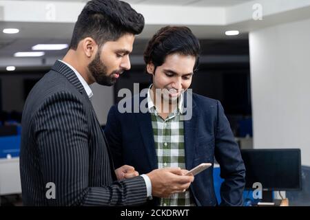 Portrait of two young handsome Indian businessmen looking into phone, discussing project, corporate environment. Stock Photo
