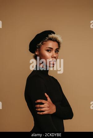 Close up portrait of african american woman standing against brown background. Woman wearing cap standing with arms crossed. Stock Photo