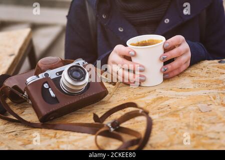 retro photo camera in leather case on a table and womans hands holding coffee cup bought from food truck in autumnal cold city park Stock Photo
