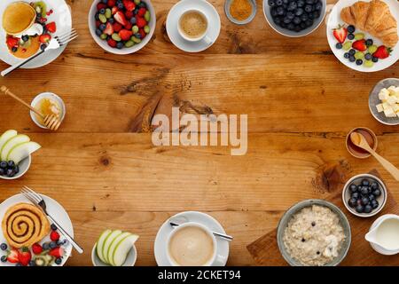 A border of delicious breakfast plates, pancakes and pasteries, bowls of fruit and porridge, and cups of coffee, on a rustic wooden background with sp Stock Photo