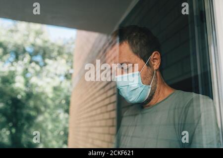 Worried man with protective face mask in self-isolation home quarantine during coronavirus outbreak, portrait of adult male person behind window Stock Photo