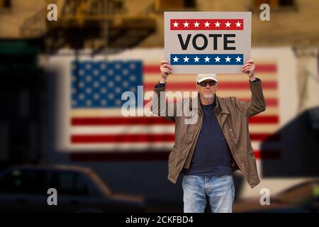 Man with cap blue jeans and  sunglasses holding a cardboard sign text VOTE above his head with american stars and stripes flag on a wall in the backgr Stock Photo