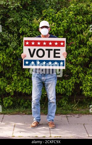 Man in full figure with cap blue jeans and medical mask holding a cardboard sign text VOTE. American Presidential patriotic background election day. Stock Photo