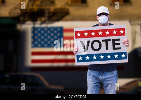 Man with cap blue jeans and medical mask holding a cardboard sign text VOTE with american stars and stripes flag on a wall in the background. American Stock Photo