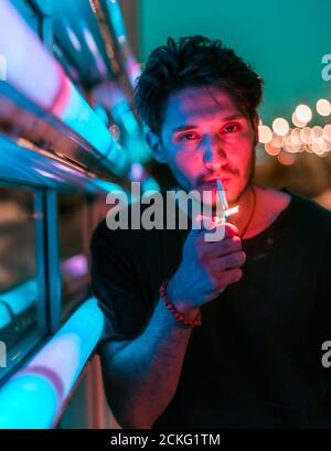 a young handsome man leaning on the neon display at night and lighting a cigarette Stock Photo