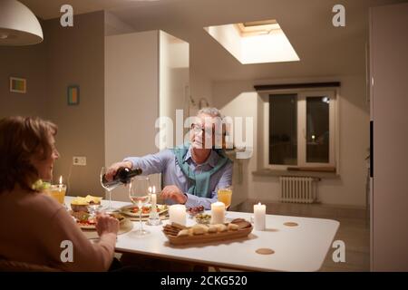 Senior man pouring the red wine into the glasses while having dinner with his wife at the table in the room Stock Photo