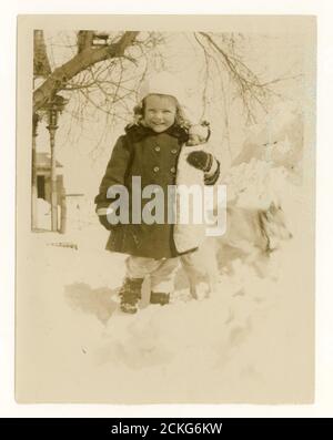 1950's photograph of a pretty happy smiling young girl standing in snow holding a doll, wintertime, U.K. Stock Photo
