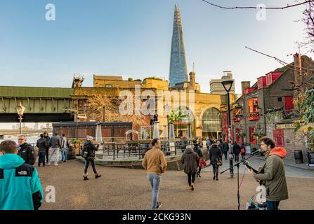 People walking along Bank Side in the late afternoon sunshine, as a busker plays near the Anchor Bankside pub with The Shard in the distance, London Stock Photo