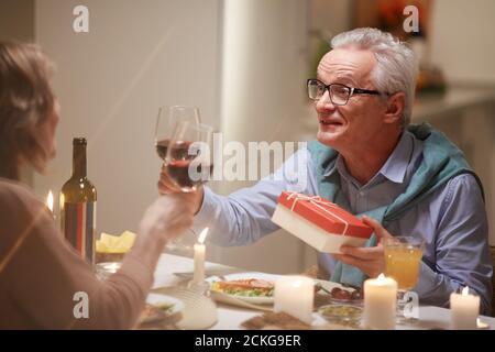 Senior man in eyeglasses holding gift box and talking to woman while they drinking wine during date Stock Photo