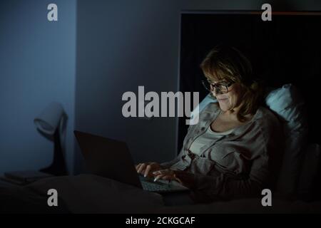 Mature woman lying on bed and typing on laptop computer in the evening in the bedroom Stock Photo