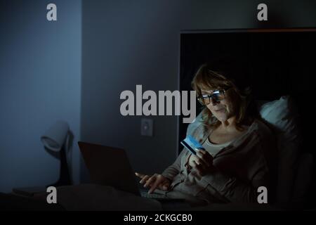 Mature woman in eyeglasses lying on bed and using laptop for online shopping in the bedroom Stock Photo