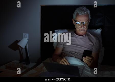Senior man in eyeglasses lying on bed and using his mobile phone and laptop in online work he working till late Stock Photo