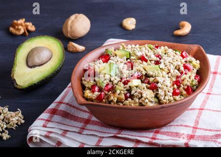 Salad of germinated buckwheat, avocado, walnut and pomegranate seeds in clay plate on black wooden background. Side view. Stock Photo