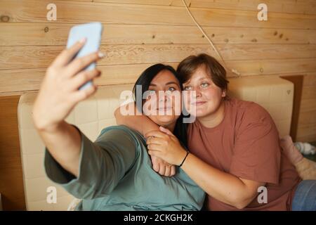 Two girlfriends embracing each other and making selfie on the phone while resting on bed Stock Photo