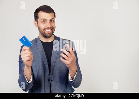 Young businessman man with a beard in a jacket, Handsome man using a credit card to pay online using a smartphone. Portrait of a man on a gray background. Stock Photo