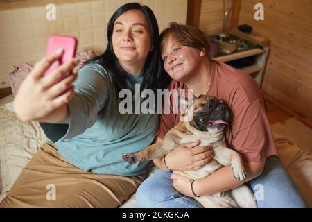 Two overweight women sitting on bed and making selfie portrait on mobile phone with their pet Stock Photo