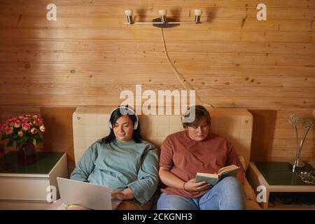 Two young overweight women relaxing on bed with laptop and book in the bedroom Stock Photo