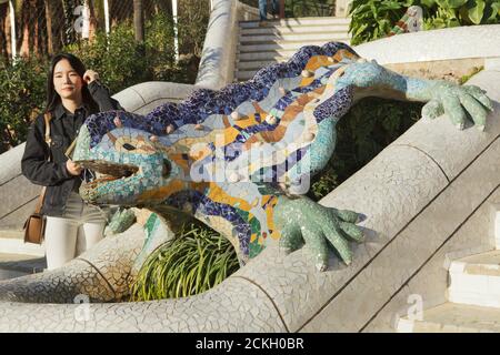 Asian tourist poses next to the trencadís mosaic salamander statue on the main staircase in the Park Güell designed by Catalan modernist architect Antoni Gaudí and built between 1900 and 1914 in Barcelona, Catalonia, Spain. Stock Photo