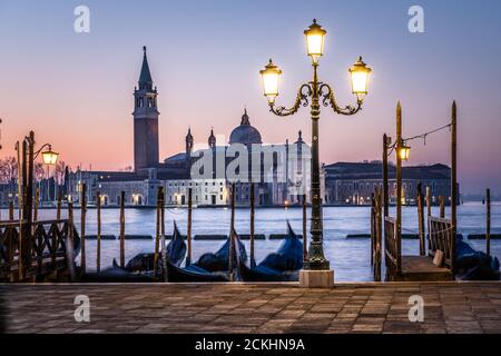 A lamp on the Riva degli Schiavoni waterfront with docked gondolas and San Giorgio Maggiore basilica in the background during a sunsrise in Venice, It Stock Photo