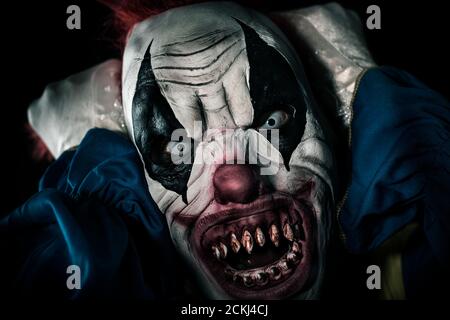 closeup of a scary evil clown with frightening white eyes and bloody teeth, pulling his red hair while staring at the observer with a threatening look Stock Photo