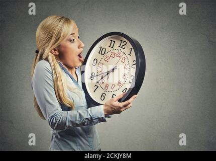 Closeup portrait woman, worker, holding clock looking anxiously, pressured by lack, running out of time, isolated on gray wall background. Human face Stock Photo