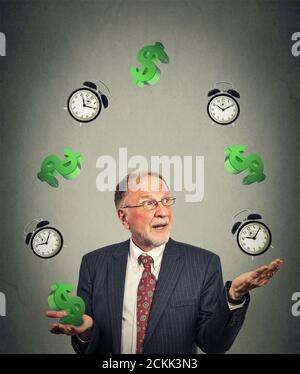 Senior business man in suit juggling multiple alarm clocks and dollar sings on gray office wall background. Time is money concept Stock Photo