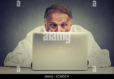 Closeup portrait elderly surprised man sitting at table working on laptop computer looking carefully at screen isolated on gray office wall background Stock Photo