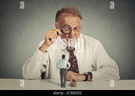 Curious corporate businessman skeptically meeting looking through magnifying glass at small employee standing on table isolated grey office wall backg Stock Photo