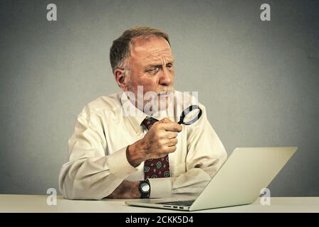 Curious. Portrait senior man investigator working looking through magnifying glass at laptop screen isolated on gray wall background. Human face expre Stock Photo