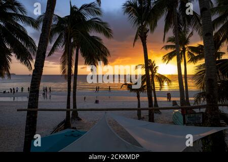 (Selective focus) Stunning view of a beautiful sunset with palm trees and the silhouette of some people relaxing on the beach. Boracay, Philippines. Stock Photo