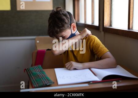 Boy wearing face mask sneezing while sitting on his desk at school Stock Photo