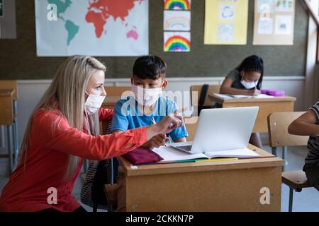 Female teacher and boy wearing face masks using laptop in class at school Stock Photo