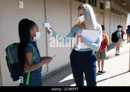 Female teacher measuring temperature of a girl wearing face mask at school Stock Photo