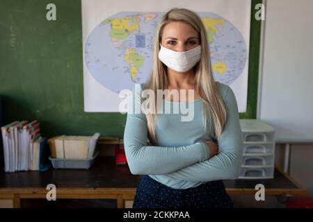 Portrait of female teacher wearing face mask standing with her arms crossed in class at school Stock Photo